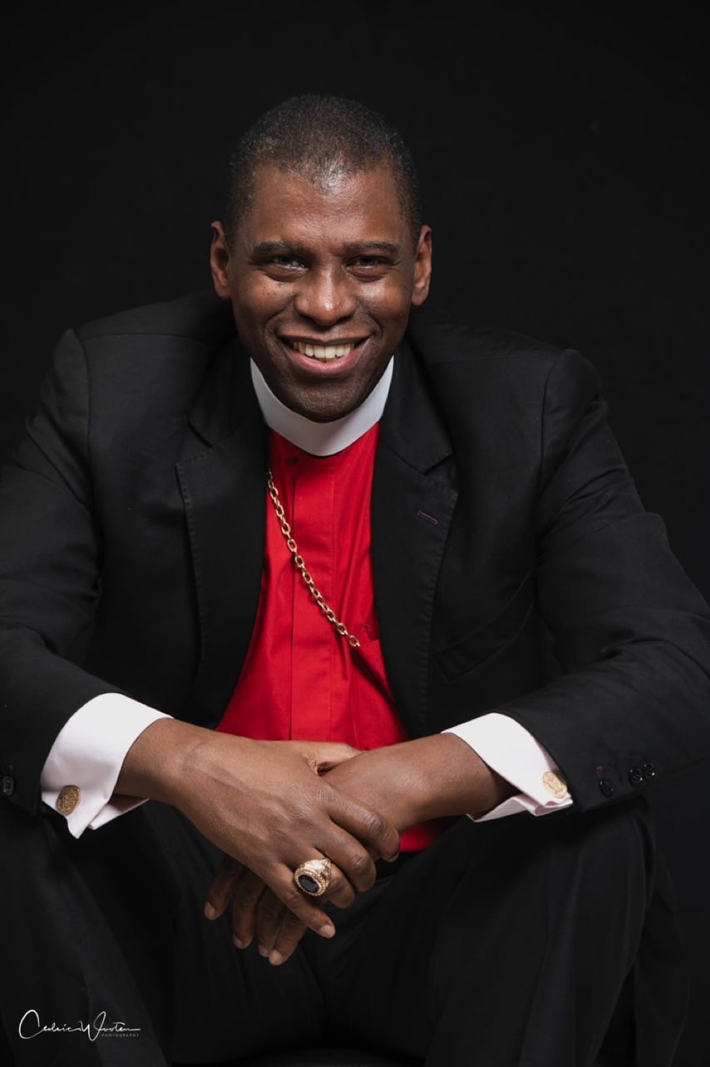 Bishop H. Curtis Douglas smiling, seated, wearing a black suit and red clerical shirt